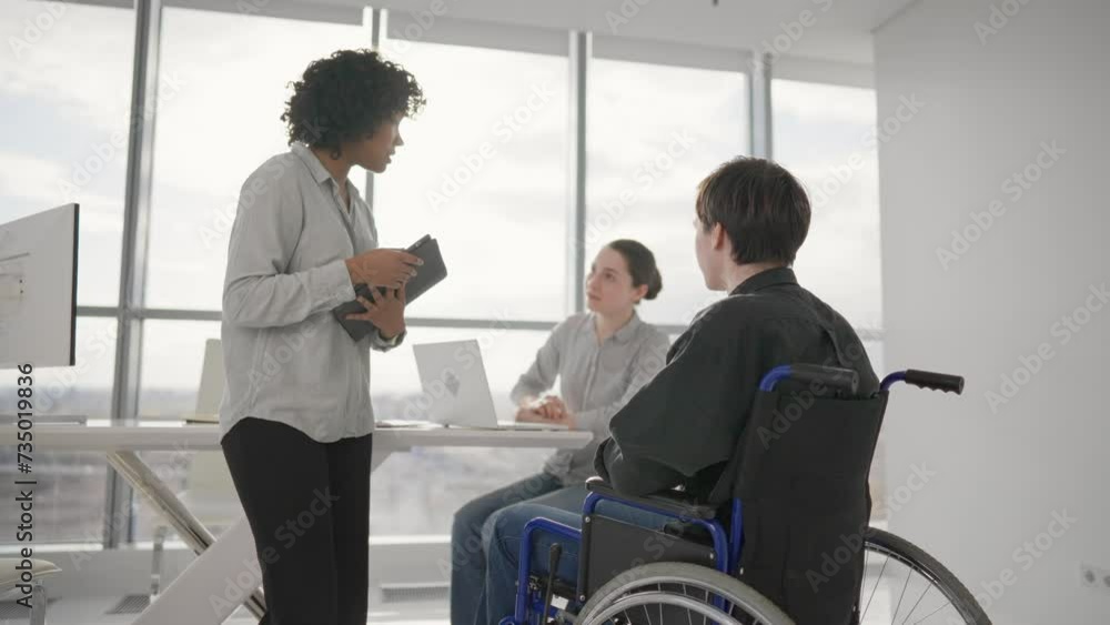 Paralyzed man works with colleagues. Guy in wheelchair has spine trauma discussing project in office on meeting with multiethnic team women colleagues. Teamwork brainstorming, special needs concept. Paralyzed man works with colleagues. Guy in wheelchair has spine trauma discussing project in office on meeting with multiethnic team women colleagues. Teamwork brainstorming, special needs concept.