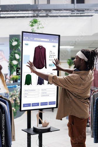 Online store buyer looks for clothes on touch screen board in clothing store, shopping for fashion items from self ordering kiosk display. Male client using interactive digital monitor. photo