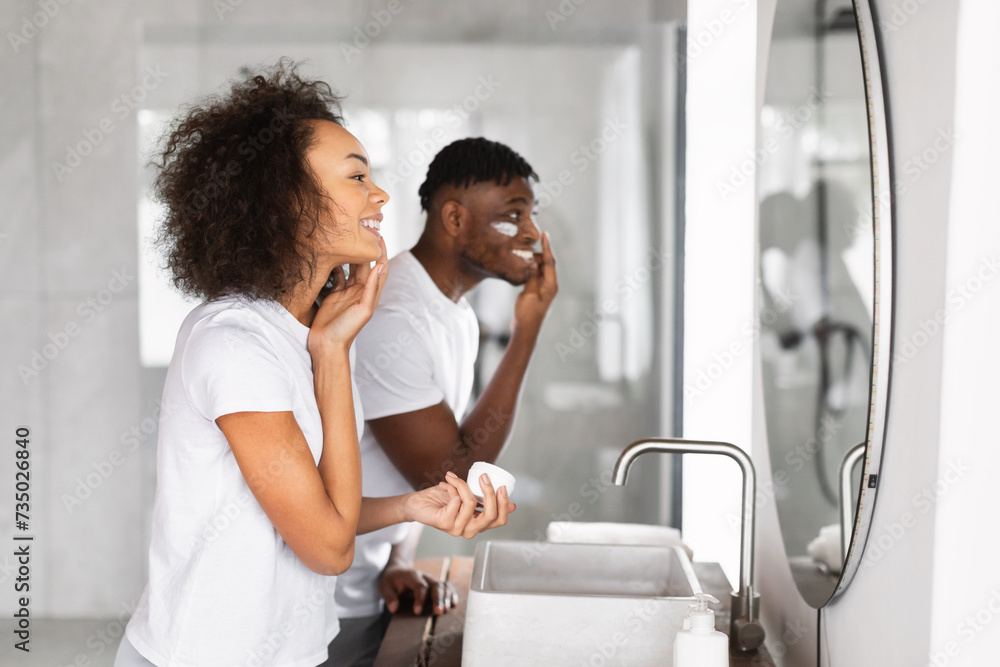 Loving black couple happily applying moisturizer near mirror in bathroom Loving black couple happily applying moisturizer near mirror in bathroom
