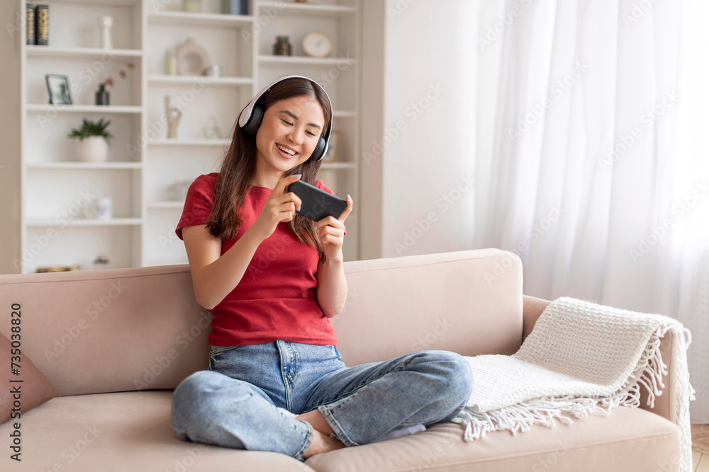 Cheerful korean woman playing video games on smartphone while relaxing on couch Cheerful korean woman playing video games on smartphone while relaxing on couch