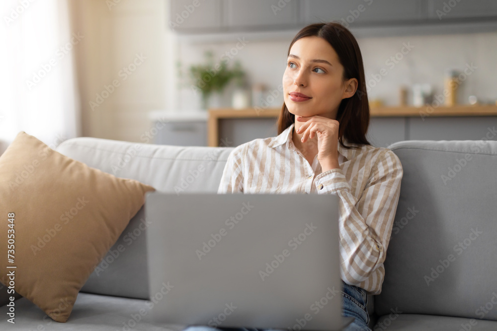 Thoughtful young woman sitting with laptop and looking aside at free space Thoughtful young woman sitting with laptop and looking aside at free space