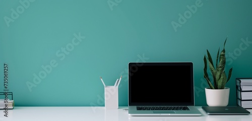 Office setup,laptop, papers, pencil holder on white table; teal backdrop. photo