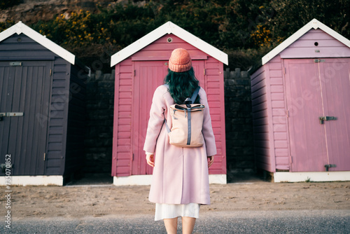 Back view stylish hipster woman with color hair in total pink outfit and backpack looking at wooden beach huts. Off season Travel concept. Seasonal street fashion. Barbiecore style. Simple pleasures. photo