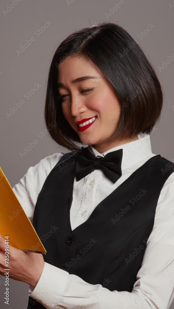 Vertical Video Asian receptionist reading a literature book over grey background, enjoying lecture hobby with a novel story in studio. Hotel concierge employee reads a fiction tale for knowledge Vertical Video Asian receptionist reading a literature book over grey background, enjoying lecture hobby with a novel story in studio. Hotel concierge employee reads a fiction tale for knowledge