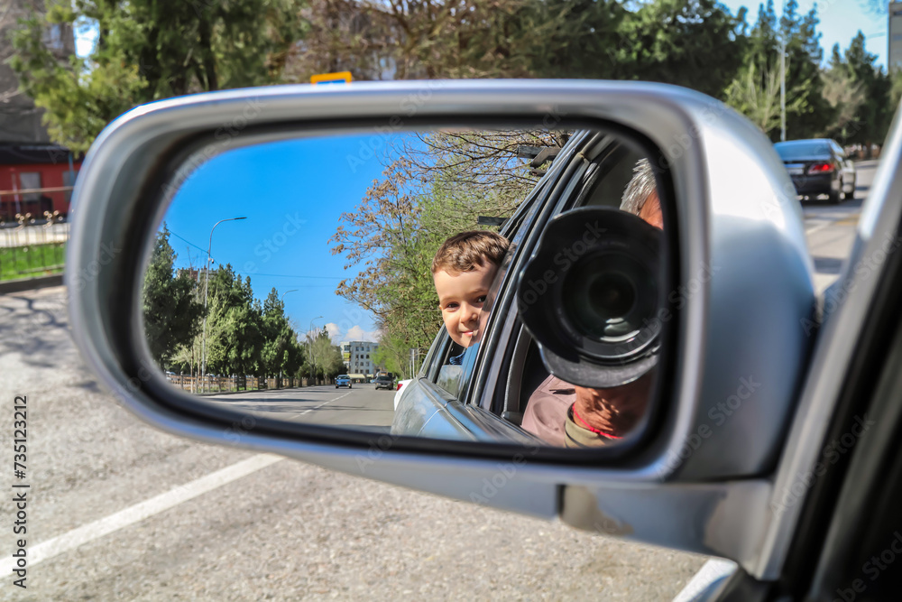 Selective focus of happy adorable little kid looking in the side mirror of a car.Man takes a picture from the camera in the mirror, reflection of the image in the car.