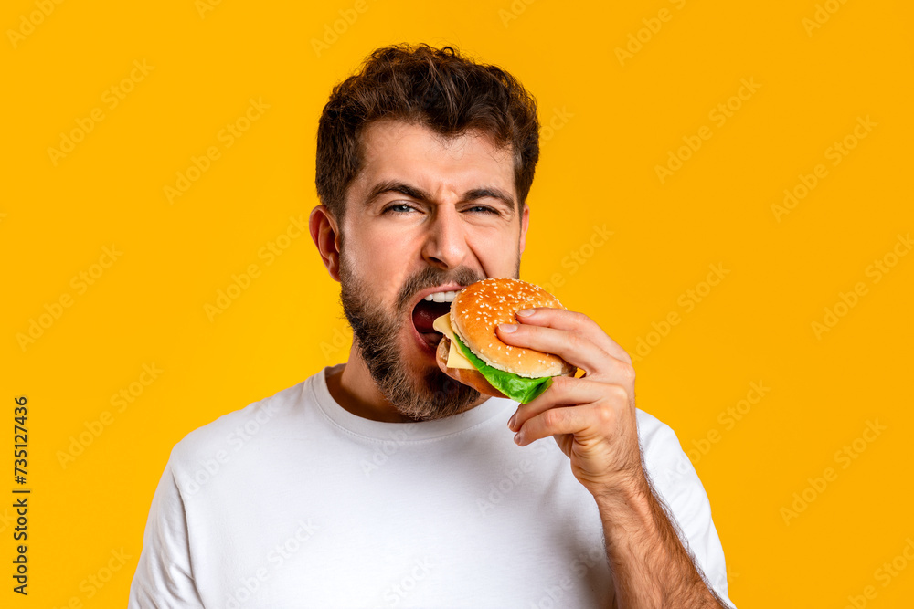 Hungry Guy Taking Bite Of Burger On Yellow Backdrop, Closeup Hungry Guy Taking Bite Of Burger On Yellow Backdrop, Closeup