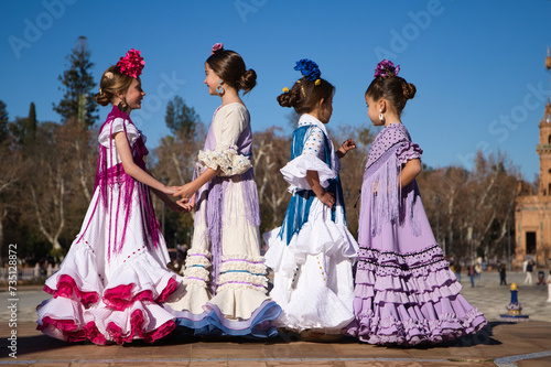 Four little girls dancing flamenco dressed in typical flamenco dress talk to each other in a famous plaza in sevilla, spain. In the background an old square tower.