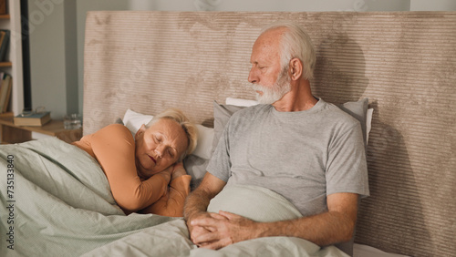 Senior man sitting on bed in his bedroom watching his wife sleep
