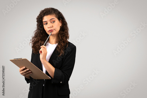 Pensive european businesswoman holding clipboard and pen to her chin, looking at free space