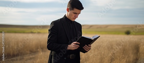 Side view of young preacher in black apparel looking in open Holy Bible while reading verses from one of canonic Gospels outdoors. Creative Banner. Copyspace image photo
