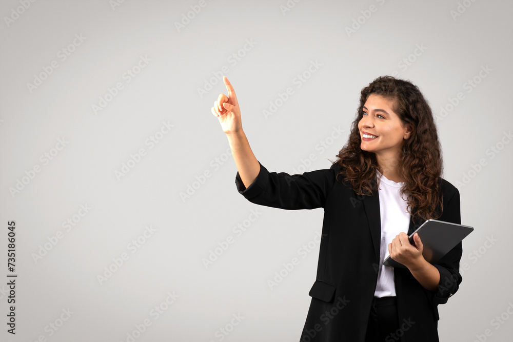 Happy young businesswoman in suit holding tablet and touching something invisible or interact with virtual interface