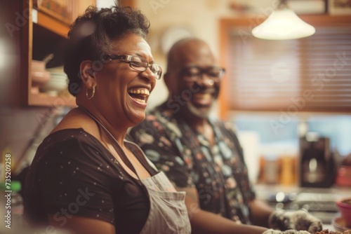 A couple share a moment of joy, their faces radiating with laughter as they stand in their cozy kitchen, surrounded by warm walls and the woman's stylish glasses