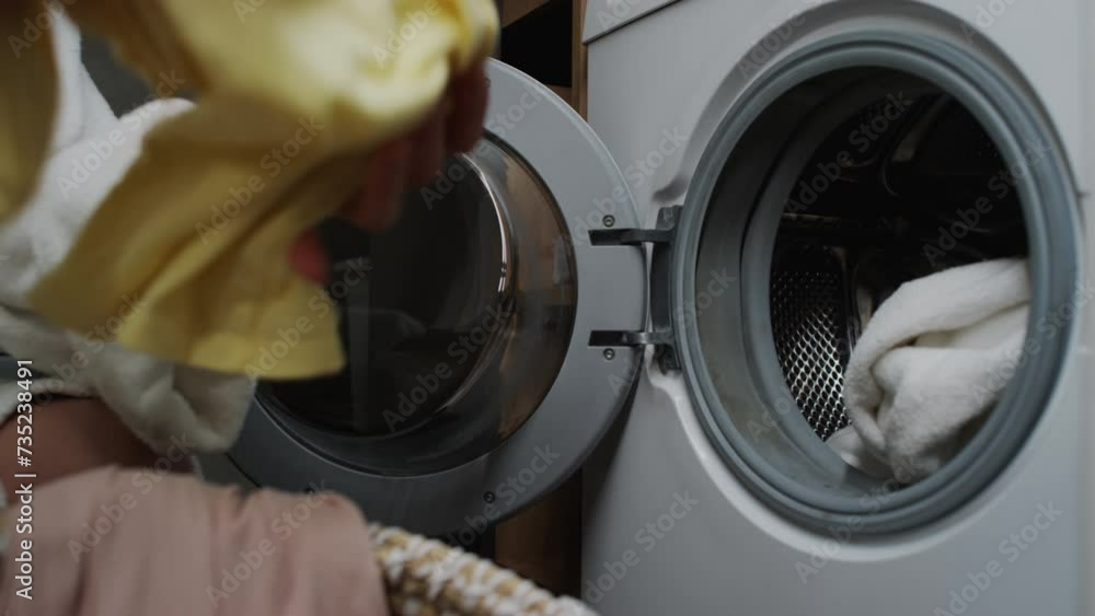 Close-up shot of hands of anonymous young woman in white bathrobe taking dirty clothes out of laundry basket, loading into washing machine and closing door Close-up shot of hands of anonymous young woman in white bathrobe taking dirty clothes out of laundry basket, loading into washing machine and closing door