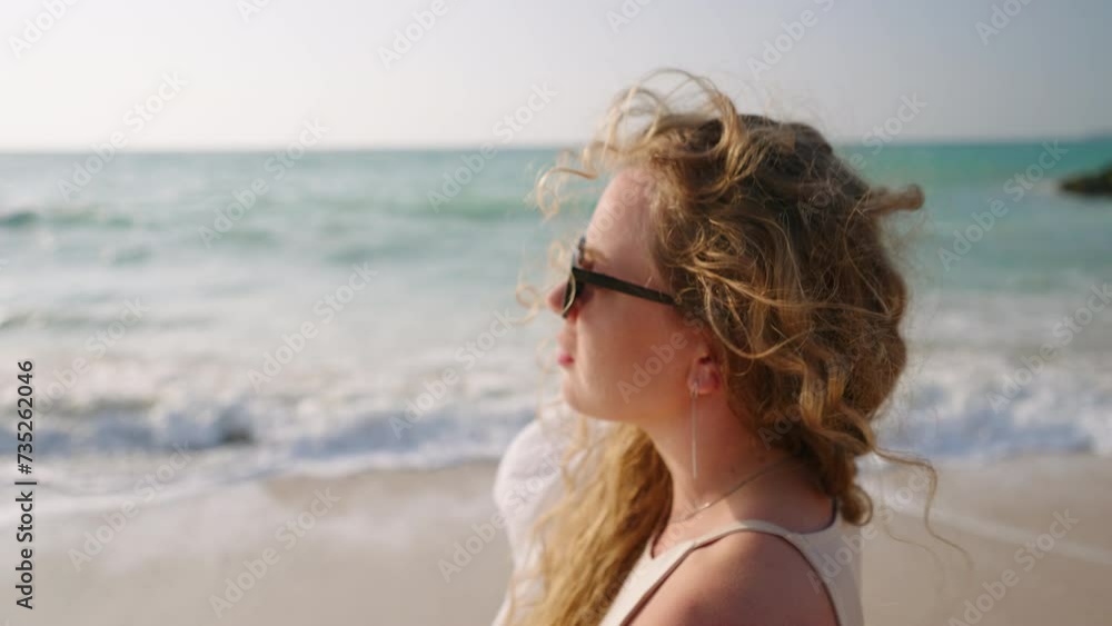 Pretty curly girl in sunglasses walking, touching hair at coast, sea surf. Young model posing, taking sun bath at ocean shore. Attractive woman enjoying summer vacation at the seaside. Close-up. Pretty curly girl in sunglasses walking, touching hair at coast, sea surf. Young model posing, taking sun bath at ocean shore. Attractive woman enjoying summer vacation at the seaside. Close-up.