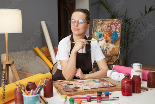 Craft education studio. Professional artist workshop. Artwork inspiration session. Pensive Caucasian woman painter wearing brown apron in art studio drawing picture
