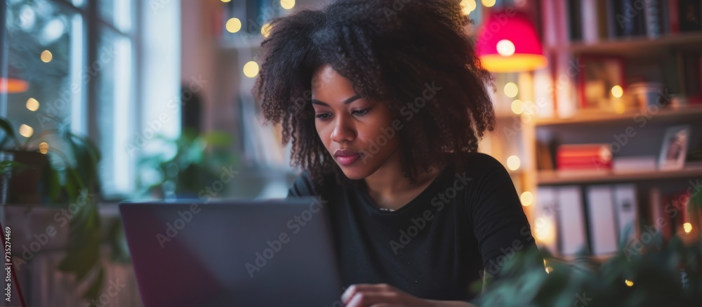 A woman is seated at a table in a room, engrossed in using her laptop. She is having fun while attending a virtual event. - obrazy, fototapety, plakaty 