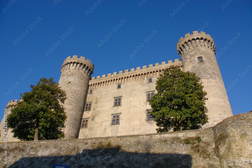 Castello di Bracciano, tall towers, tree at base, bright sky.