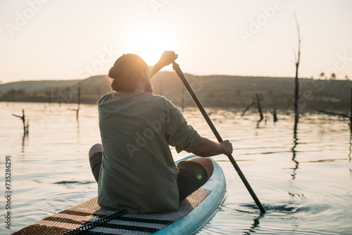 A man is surfing on a sap board on calm water at sunset.