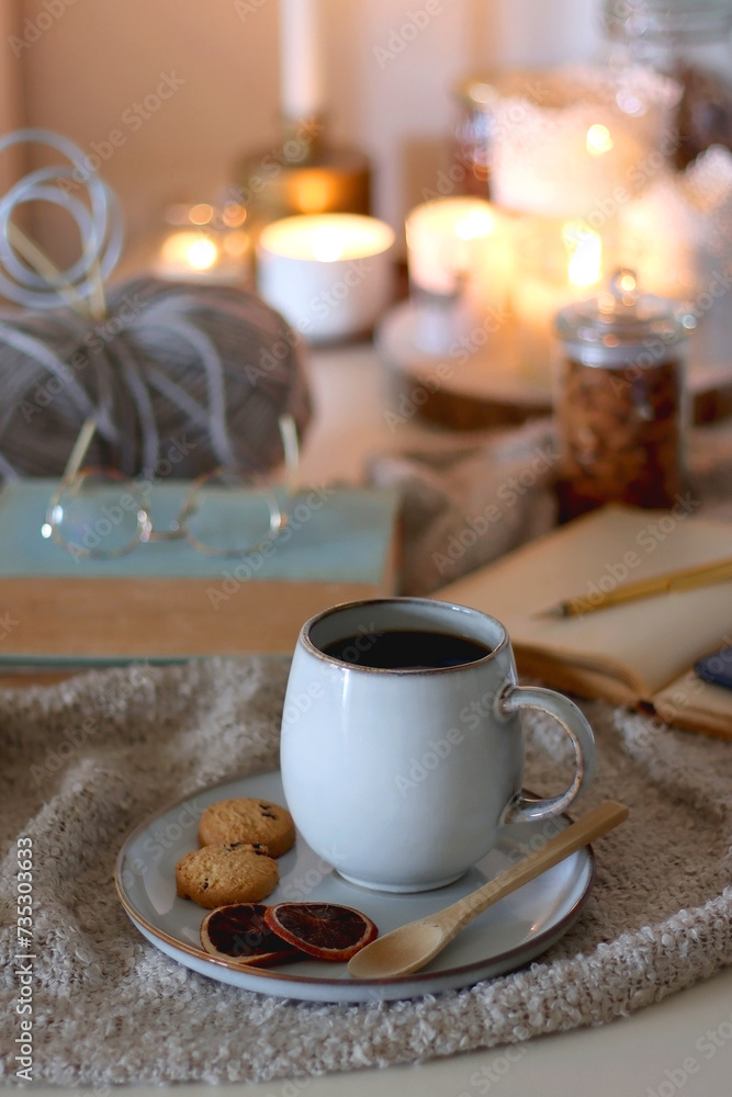 Cup of tea or coffee, cookies, books, glasses, e-reader, almonds, ball of yarn, knitting needles and lit candles on the table. Hygge at home. Selective focus.