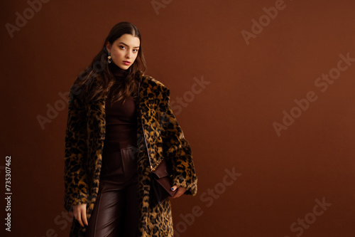 Fashionable confident woman wearing trendy faux fur leopard print coat, turtleneck, leather pants, posing on brown background. Studio fashion portrait. Copy, empty space for text