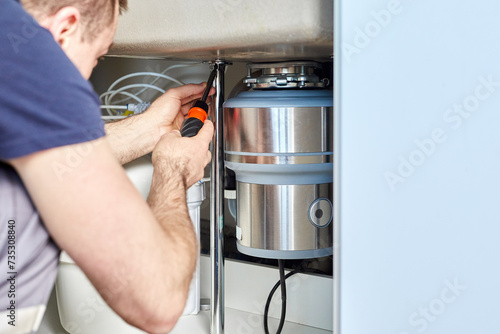 A plumber with gloved hands installs a household waste shredder for the kitchen sink. A technician installs or repairs a household waste shredder in the kitchen.