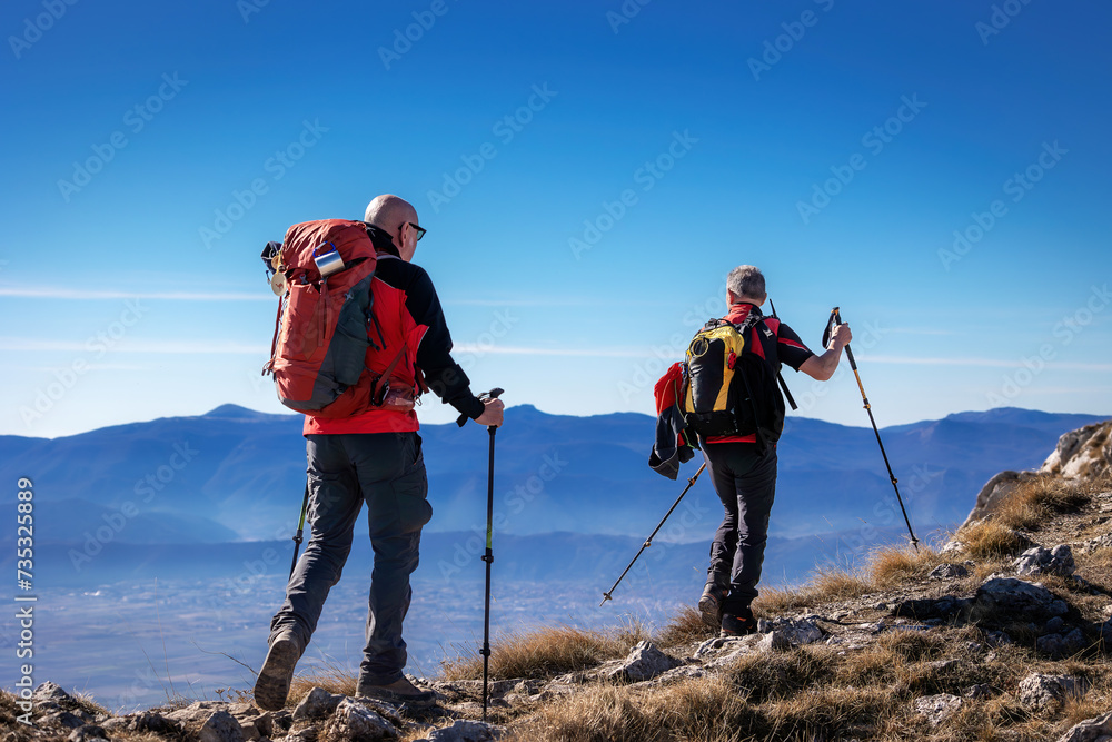 Two hikers reach the summit of the mountain, helping themselves to climb with trekking poles. In the background the plain surrounded by mountains.