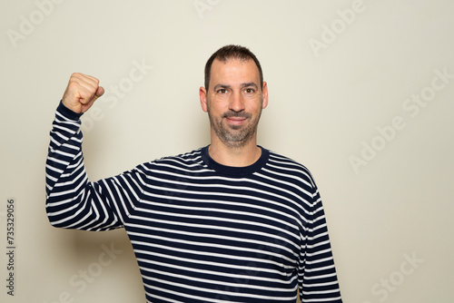 Hispanic man with a beard of 40 years old showing biceps believing that his training has paid off, the poor deluded man does not realize his terrible physical condition. Isolated beige background. photo