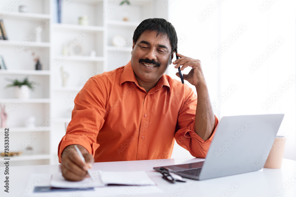 Smiling mature indian man working on laptop, have phone conversation Smiling mature indian man working on laptop, have phone conversation