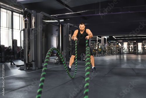 Crossfit training. Motivated muscular man exercising with battle ropes at gym
