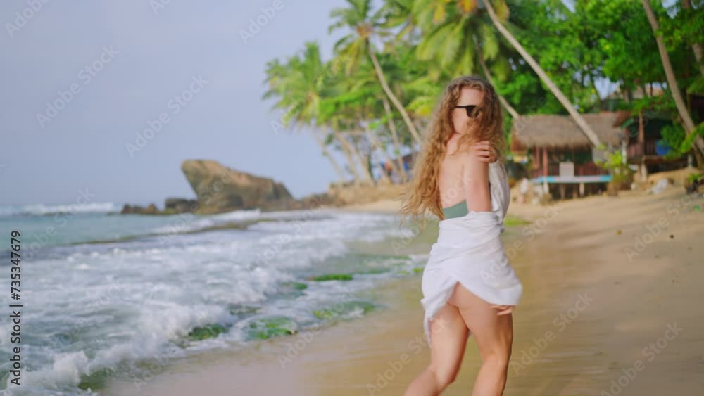 Curly-haired woman in green swimsuit, white sarong, strolls along sandy shore, invites viewers to tropical beach getaway. Smiling seductively, suggests joining under palm trees, serene ocean backdrop. Curly-haired woman in green swimsuit, white sarong, strolls along sandy shore, invites viewers to tropical beach getaway. Smiling seductively, suggests joining under palm trees, serene ocean backdrop.