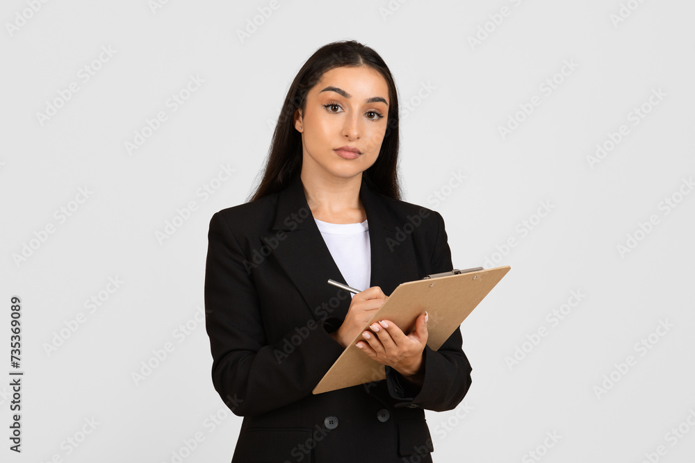 Serious businesswoman holding clipboard and writing, looking at camera Serious businesswoman holding clipboard and writing, looking at camera