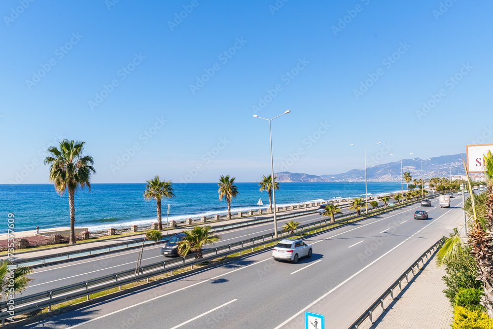 Obraz premium Highway Road with Cars Moving Next to a Blue Sea and Sandy Beach Under The Clear Sky in a Hot Sunny Summer day from a Pedestrian Stone Bridge