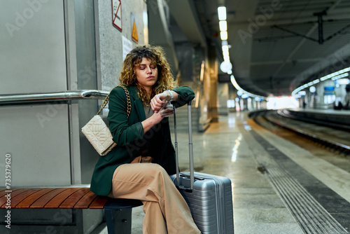 A weary businesswoman with curly hair sits, patiently awaiting her train at the station.