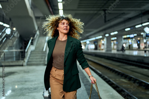 In a rush at the subway station, a curly-haired woman in her 30s dashes with a travel suitcase, capturing the thrill of travel.