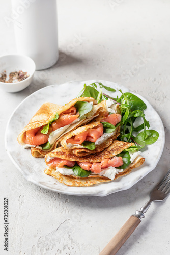 Yummy crepes or thin pancakes with smoked salmon, soft cheese and spinach on a plate on gray textured background. Pancake day, Maslenitsa