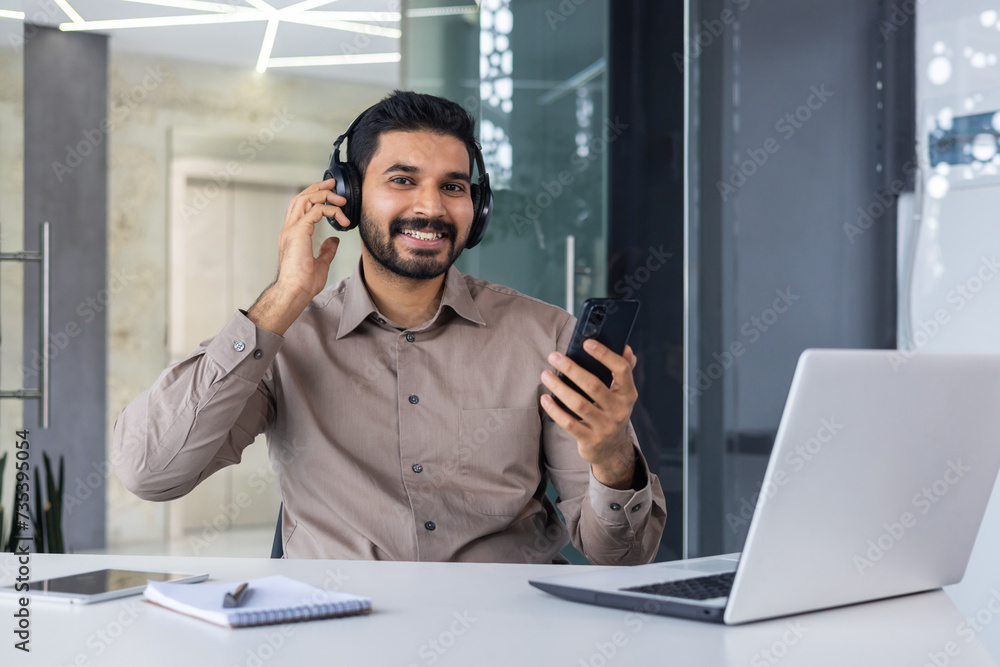 Carefree hispanic man in headphones holding cellular phone in hand while sitting by equipped desktop in office. Relaxed manager fixing wireless accessory for listening to music during workday Carefree hispanic man in headphones holding cellular phone in hand while sitting by equipped desktop in office. Relaxed manager fixing wireless accessory for listening to music during workday