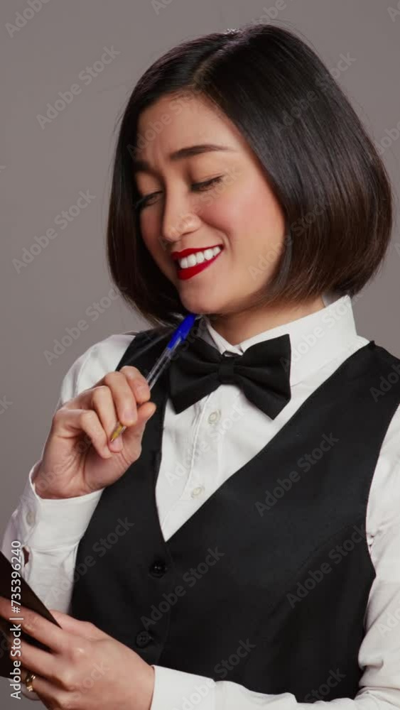 Vertical Video Asian hotel concierge creating a checklist of registrations, working on online reservations and bookings in studio. Receptionist looking at clipboard papers over grey background. Camera Vertical Video Asian hotel concierge creating a checklist of registrations, working on online reservations and bookings in studio. Receptionist looking at clipboard papers over grey background. Camera