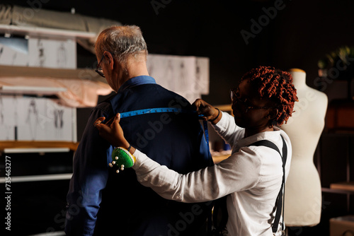 Expert seamstress sizing up stylish client proportions for upcoming luxurious bespoke costume. Elderly customer getting measurements taken by meticulous suitmaker in tailoring studio photo