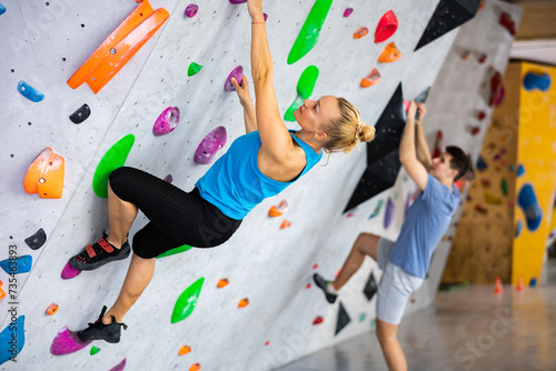 Caucasian woman exercising on wall in climbing gym during bouldering training. Young man climbing in background.