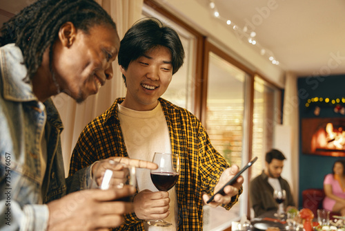 Canvas Print Two male friends drinking wine while looking on phone during festive home party