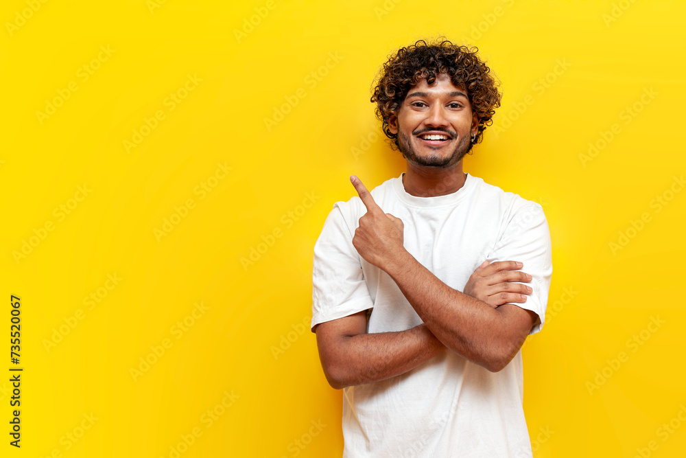 Fototapeta premium young indian man in white t-shirt pointing at copy space and smiling on yellow isolated background, curly guy showing and advertising empty space