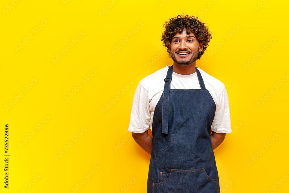 cheerful indian male waiter in an apron stands and smiles on a yellow isolated background, indian guy barista entrepreneur in uniform looks away cheerful indian male waiter in an apron stands and smiles on a yellow isolated background, indian guy barista entrepreneur in uniform looks away