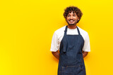 cheerful indian male waiter in an apron stands and smiles on a yellow isolated background, indian guy barista entrepreneur in uniform looks away