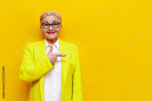 old businesswoman in glasses and formal wear pointing with hands at empty space and smiling on yellow isolated background, elderly pensioner grandmother in blazer showing and advertising copy space photo