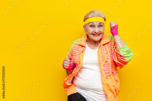 crazy funny old granny in sports colorful clothes wins and celebrates success on a yellow isolated background, elderly woman in youth hipster clothes rejoices in victory and luck and screams photo
