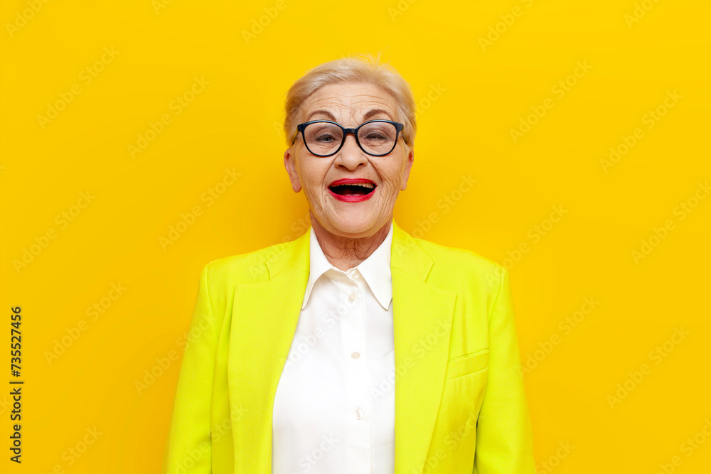 cheerful surprised old businesswoman in glasses and formal wear looking at camera with open mouth over yellow isolated background, elderly pensioner grandmother in blazer screaming in amazement