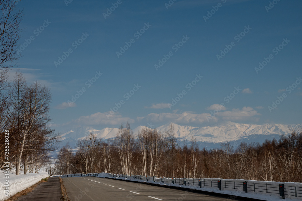 Obraz premium 雪が溶けた早春の道路と冠雪の山並み 十勝岳連峰 