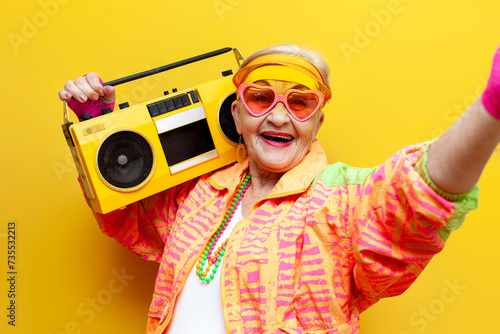 funny crazy granny in hipster clothes listening to music on tape recorder and taking selfie on yellow isolated background, elderly cool woman dancing at party and smiling