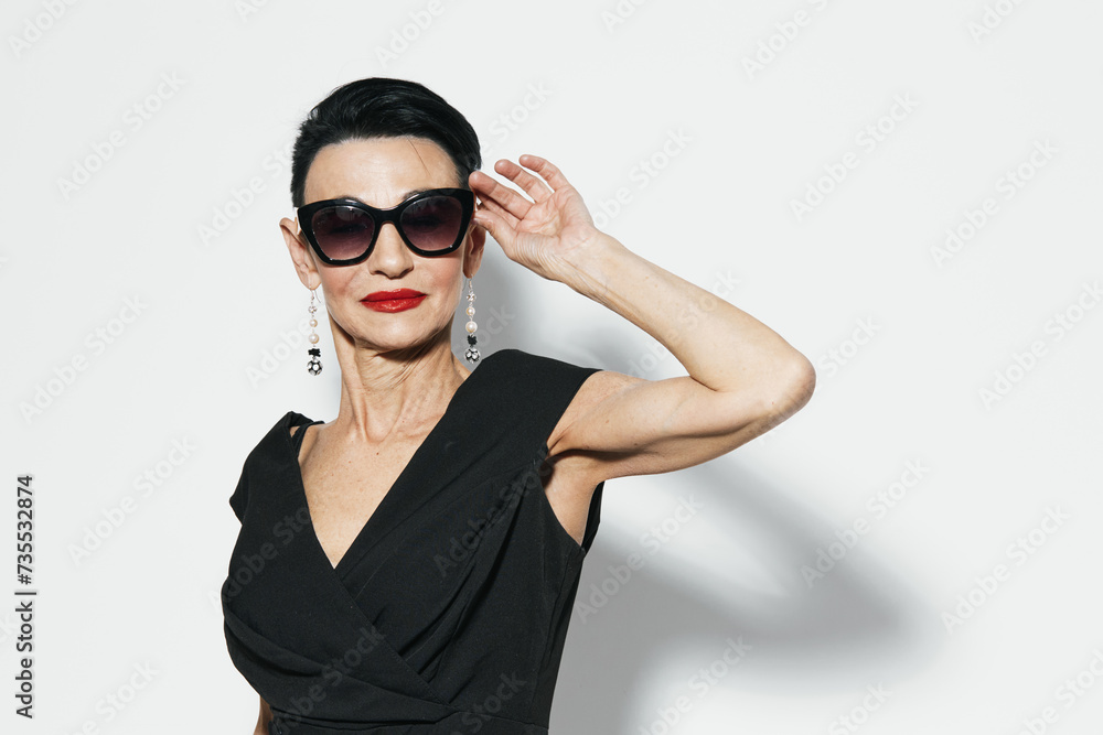 Elegant senior woman in black dress and sunglasses posing for camera in front of white wall Elegant senior woman in black dress and sunglasses posing for camera in front of white wall
