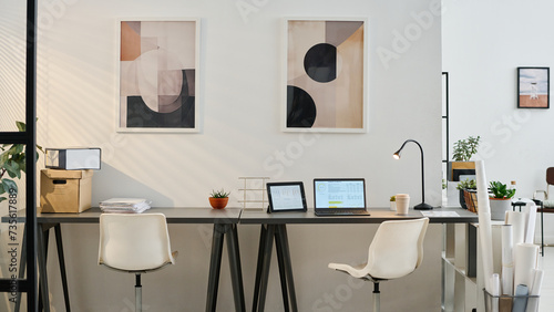 No people shot of part of modern office interior with laptop, digital tablet, pile of documents on desks and paintings on walls, copy space photo